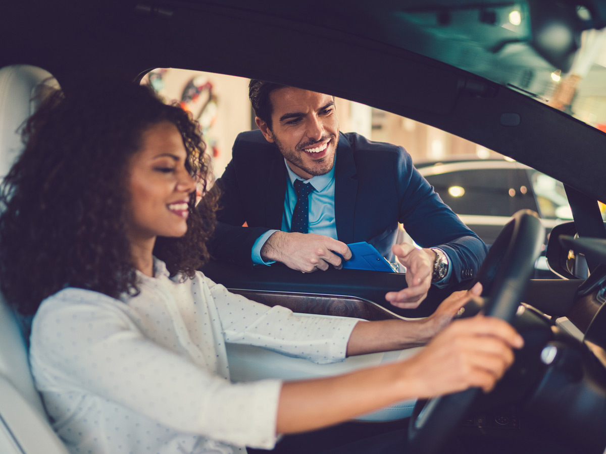 A salesman shows car safety features to a woman sitting behind the wheel of a new car