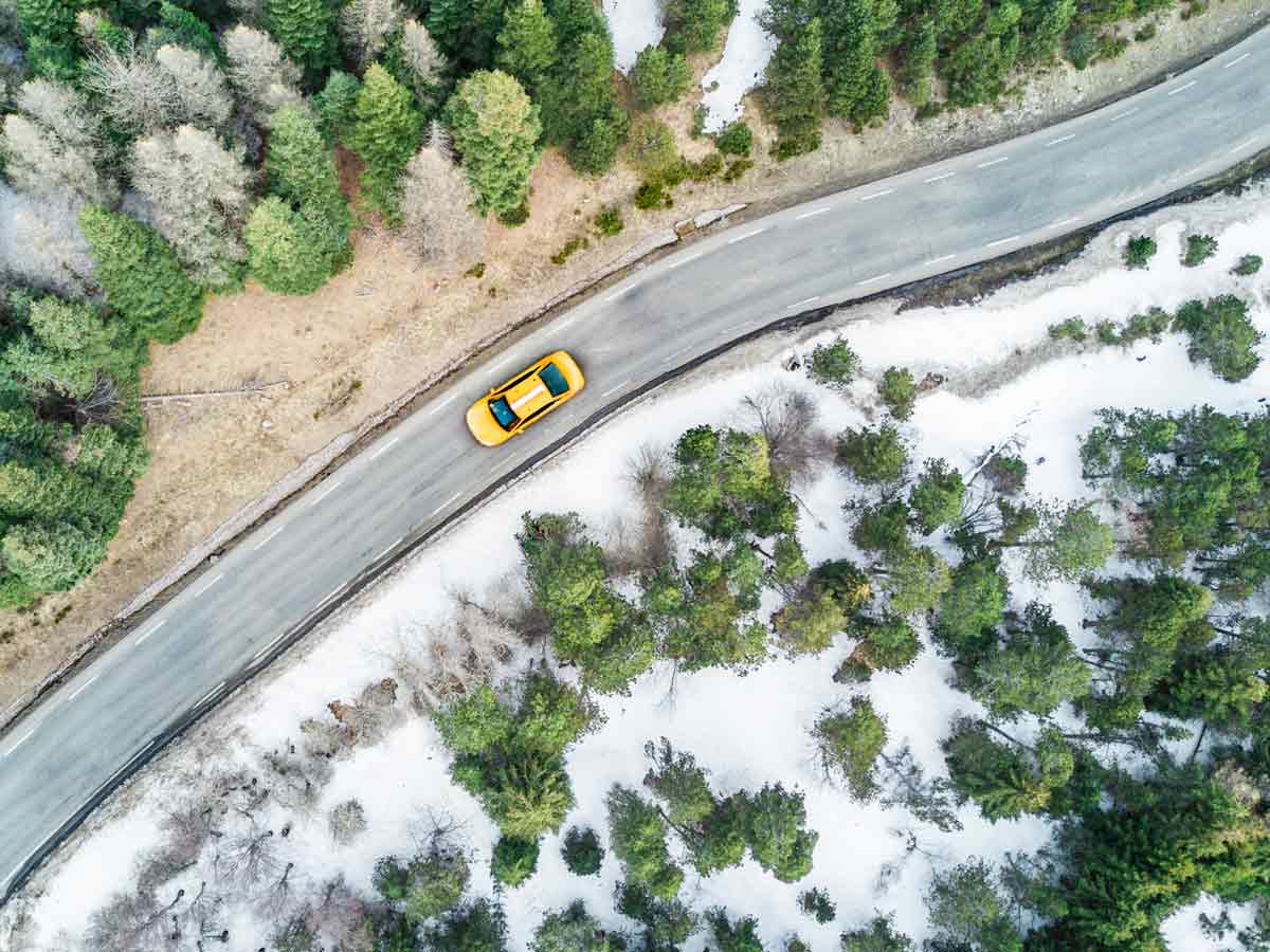 A car on a winding country road that cuts through snow covered forests.
