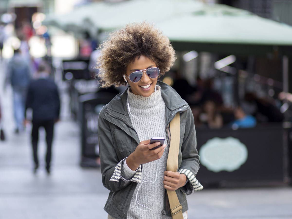 A woman walks down the street, looking at her smartphone and listening to music with earphones.