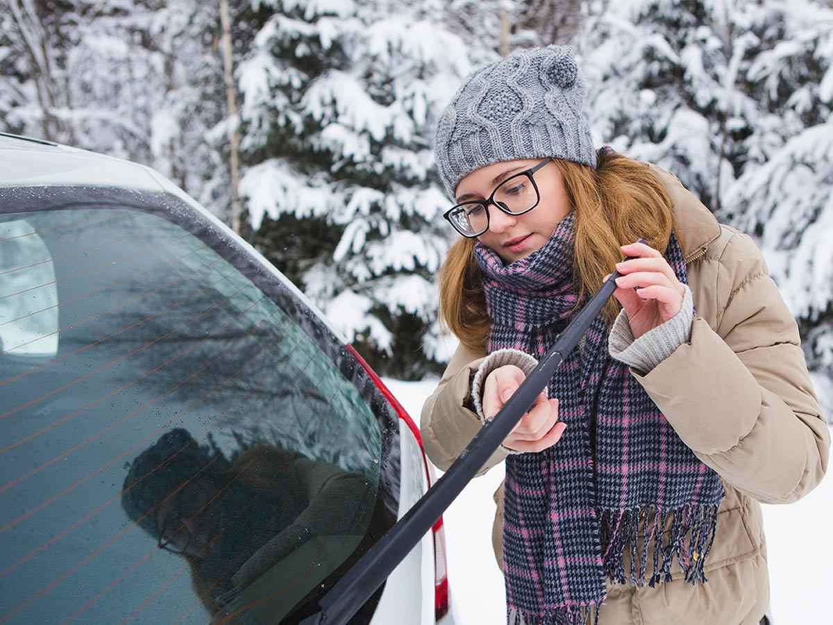 A woman in a tuque, scarf and winter coat installs winter wiper blades on her car.